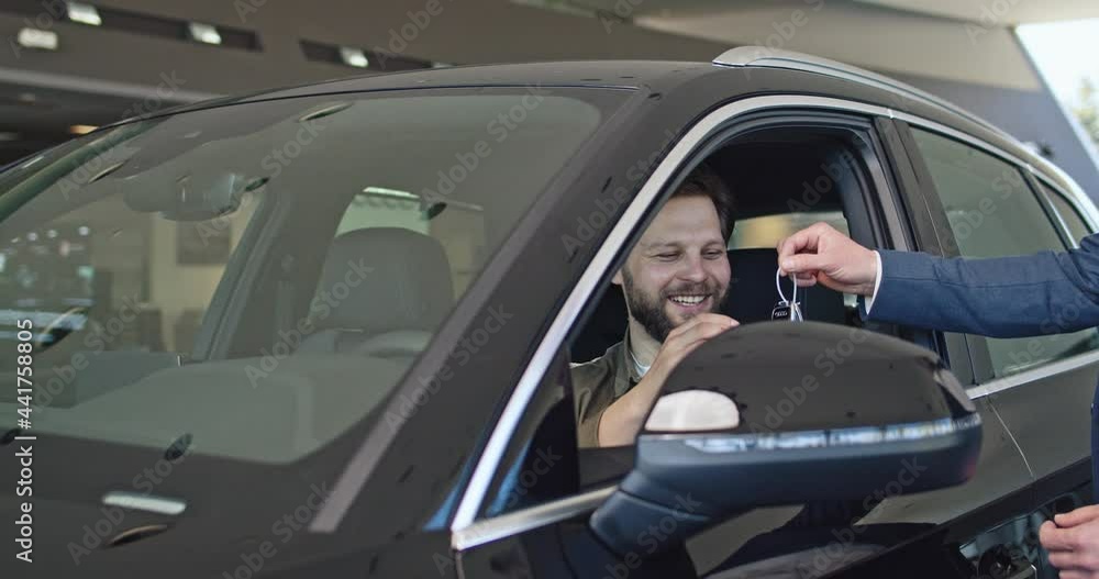 Caucasian male manager giving keys from new car to happy male client. Young caucasian man sitting in auto and feeling happiness from expensive purchase. Caucasian male manager giving keys from new car to happy male client. Young caucasian man sitting in auto and feeling happiness from expensive purchase.