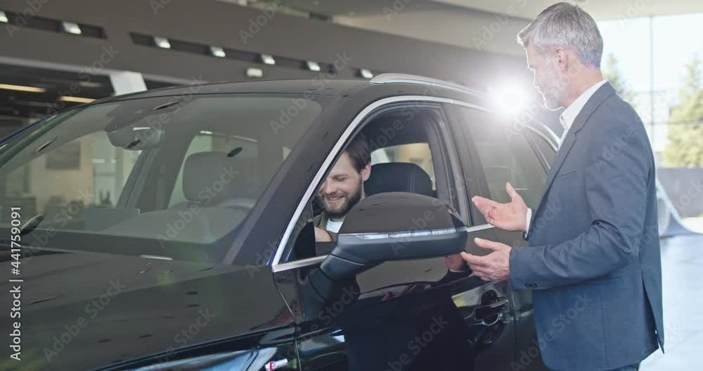 Handsome man is sitting in a car, smiling and talking to the car dealer. Trader gives key. Bearded caucasian car dealer serving customer in showroom. Handsome man is sitting in a car, smiling and talking to the car dealer. Trader gives key. Bearded caucasian car dealer serving customer in showroom.