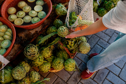 Young woman picking fresh green Italian Artichokes on farmers market. Female shopping for organic local produce vegetables. Vegan diet. Clean eating concept. Close up, copy space, background.