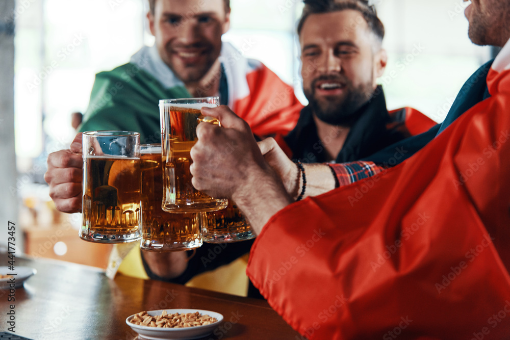 Happy young men covered in international flags toasting with beer while watching sport game in the pub