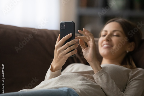Focus on smartphone in hands of relaxed beautiful woman lying on cozy couch. Addicted to modern tech pleasant happy lady typing message, web surfing information, communicating distantly or shopping.