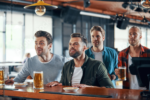 Cheering young men in casual clothing watching sport game and enjoying beer while sitting in the pub