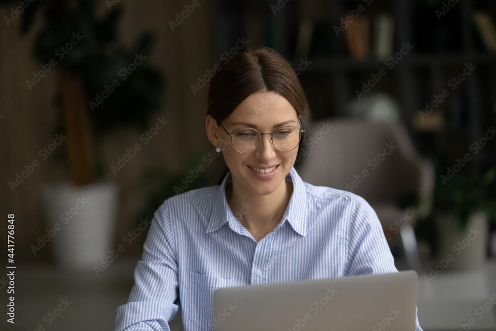 Happy young business lady employee entrepreneur in eyeglasses looking at laptop screen, enjoying working on new online project, communicating distantly, web surfing information at home office. Happy young business lady employee entrepreneur in eyeglasses looking at laptop screen, enjoying working on new online project, communicating distantly, web surfing information at home office.