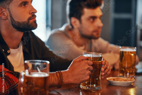 Young men in casual clothing enjoying beer while watching TV in the pub