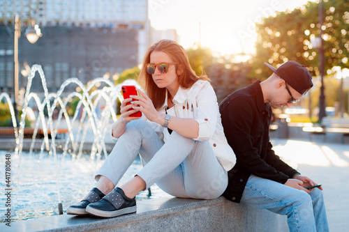 young fashionable couple, a man and a woman, uses a mobile phone in a city park near a fountain at sunset in summer.