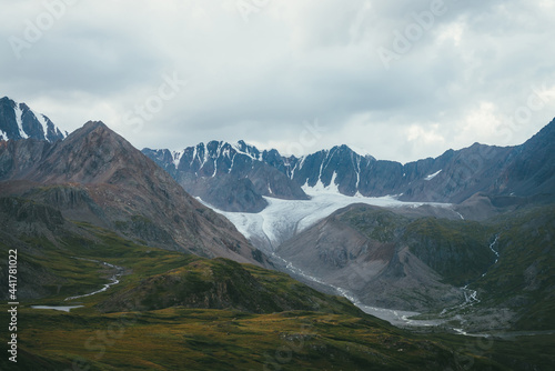 Dramatic alpine landscape with big glacier and mountain river in valley in overcast weather. Atmospheric scenery with great mountain range under cloudy sky. Beautiful mountain ridge under gray sky.