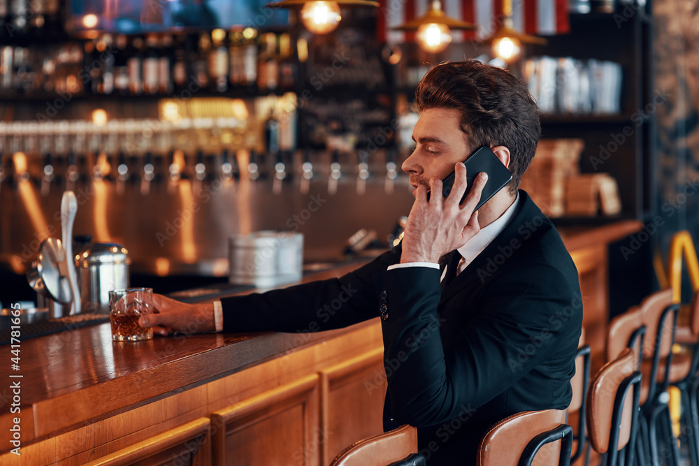 Handsome young man in full suit talking on smart phone while sitting at the bar counter in restaurant Handsome young man in full suit talking on smart phone while sitting at the bar counter in restaurant