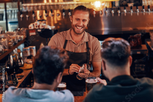 Young bartender serving beer to young men while standing at the bar counter in pub