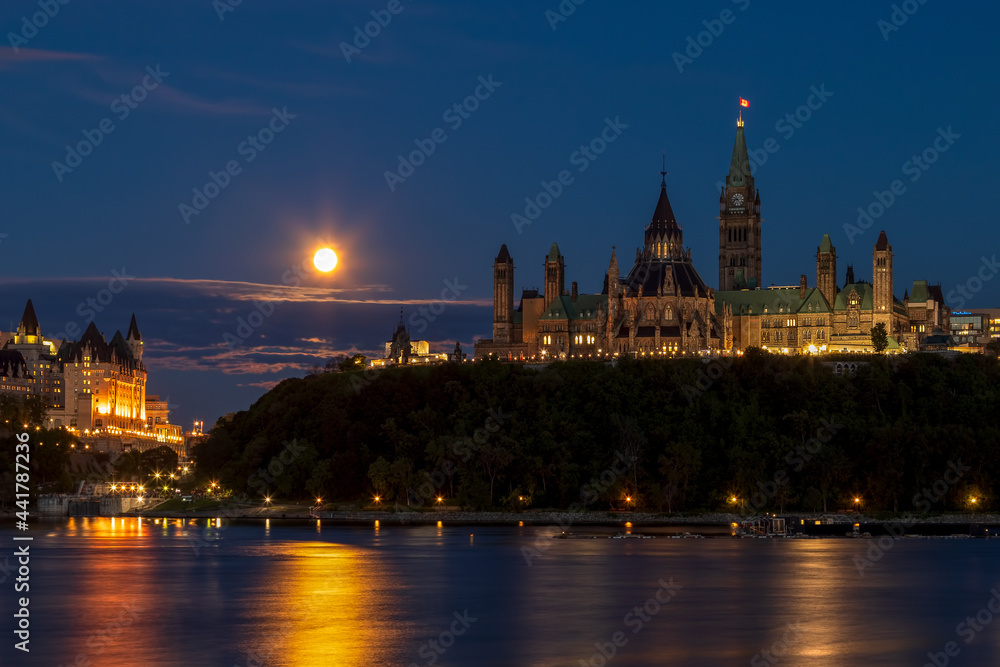 Naklejka premium Downtown Ottawa at night by the river. Full moon shining low in the blue sky. Parliament buildings and historic hotel in Ottawa, Ontario, Canada