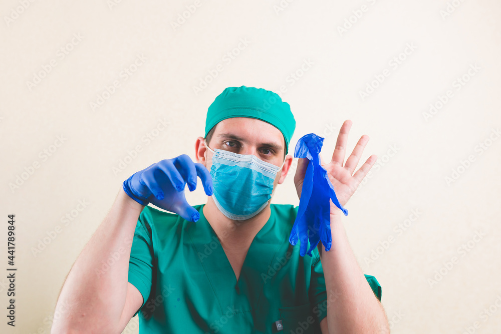 Young doctor in green surgeon suit and protective breathing mask holds blue gloves in his hands isolated. Selective focus. Young doctor in green surgeon suit and protective breathing mask holds blue gloves in his hands isolated. Selective focus.