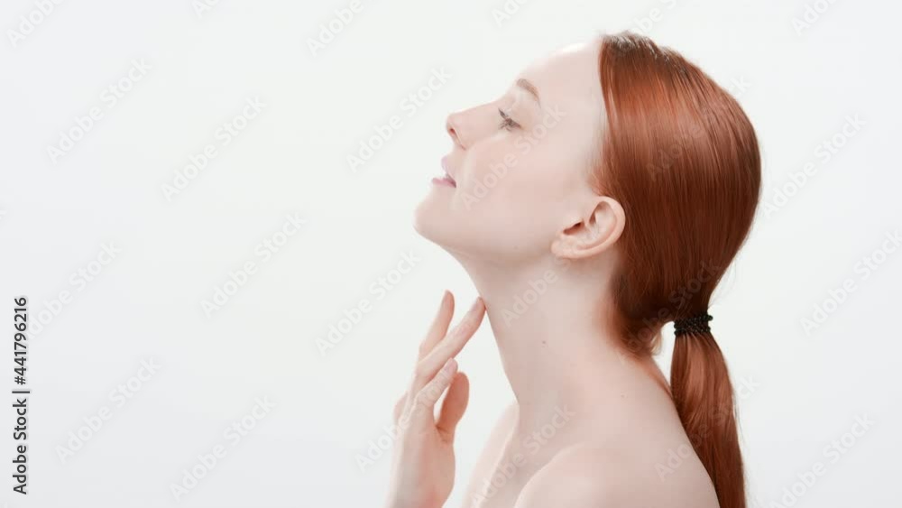 Beautiful fit European young woman with long rusty hair in ponytail touches her chin and neck standing sideways and looking up on white background | Moisturizer commercial Beautiful fit European young woman with long rusty hair in ponytail touches her chin and neck standing sideways and looking up on white background | Moisturizer commercial