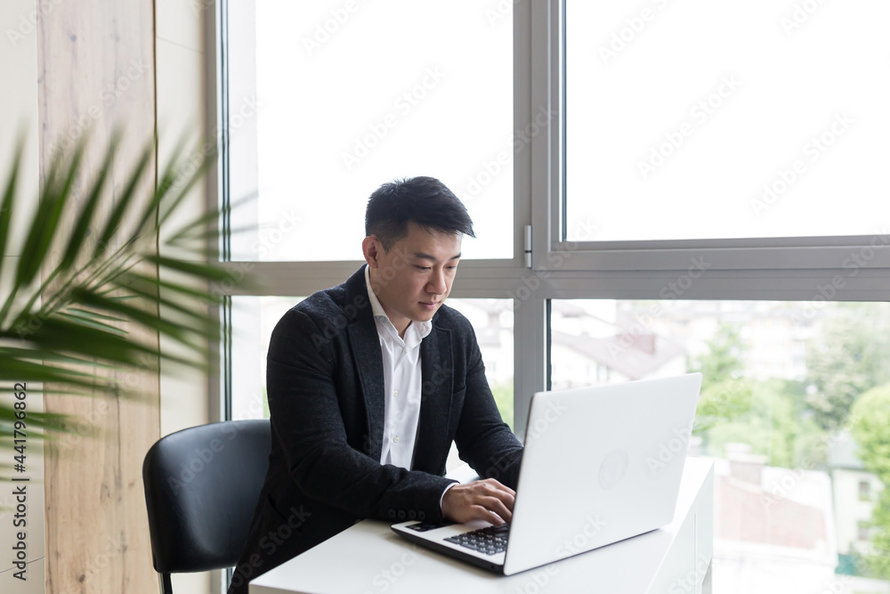 Successful Asian businessman, in a black business suit, works on a laptop, in a stylish office explains a new business strategy via video link Successful Asian businessman, in a black business suit, works on a laptop, in a stylish office explains a new business strategy via video link