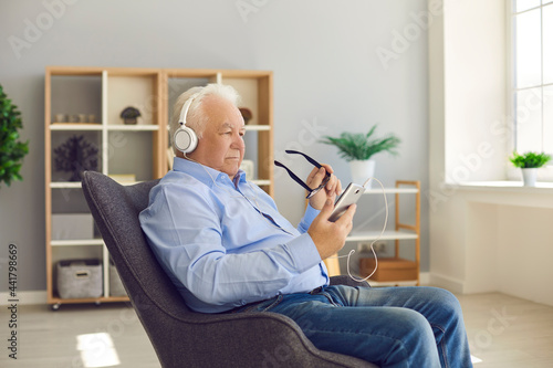 Senior man sitting in armchair at home wearing headphones and using mobile phone