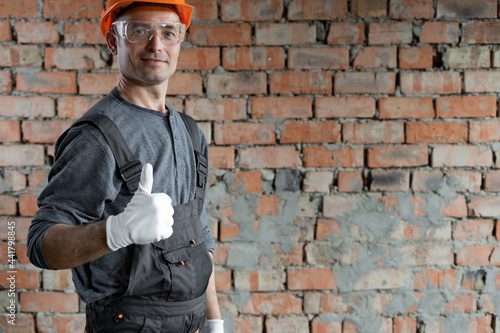 a construction engineer or an architect in a protective helmet and glasses stands indoors against a brick wall and raised his finger up shows cool. Male foreman to the left of the text space. © mtrlin