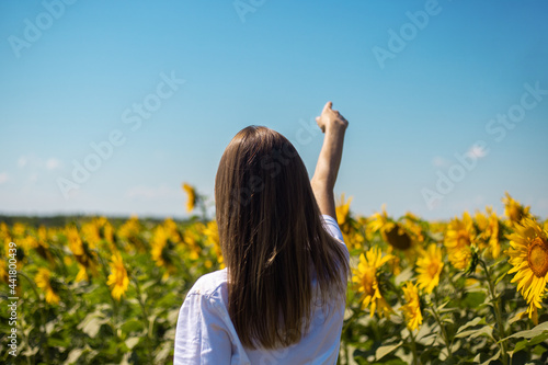 Young woman in white t-shirt points a finger at the horizon on a sunflower field on a summer sunny day