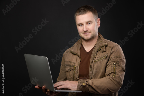Portrait of friendly positive caucasian casual man using gray laptop while standing in studio and looking at camera © yanik88