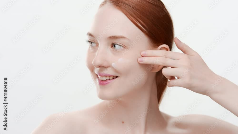 Attractive young fit Caucasian woman with long ginger hair applies nourishing cream on her cheek smiling wide on white background | Nourishing cream commercial Attractive young fit Caucasian woman with long ginger hair applies nourishing cream on her cheek smiling wide on white background | Nourishing cream commercial