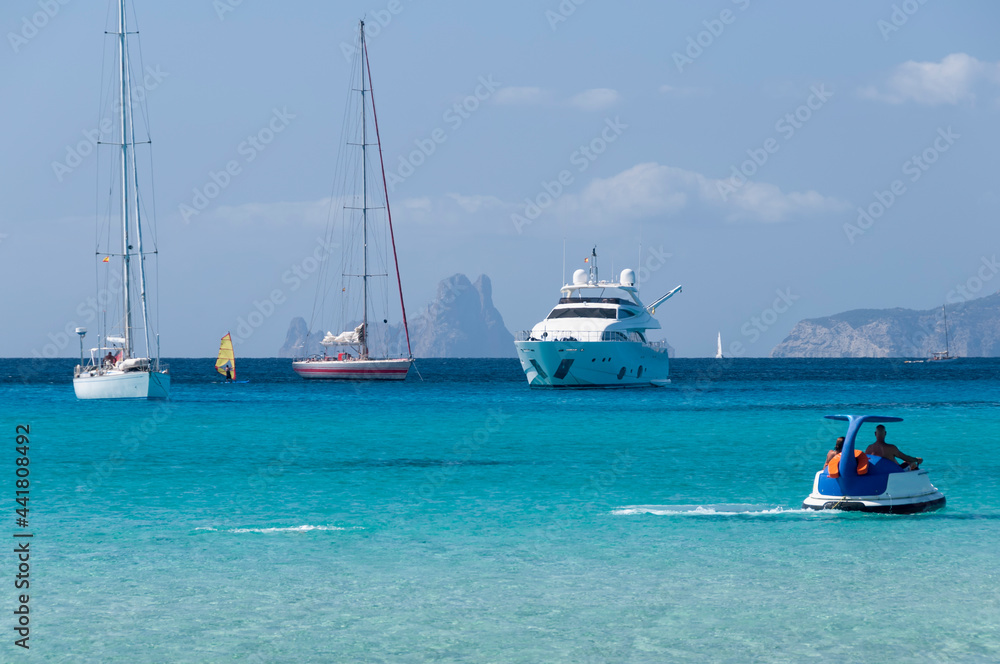 Fototapeta premium A couple of tourists sail near the beach in Formentera with a small boat. In the background luxury boats anchored in the Mediterranean Sea.