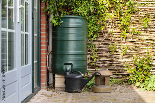 A green rain barrel to collect rainwater and reusing it to water the paints and flowers in a backyard with a wattle fence made of willow branches on a sunny day photo