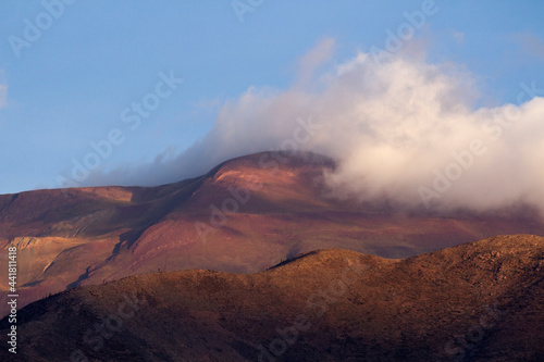 Inspiring alpine landscape. View of the brown mountain peak under a white cloud at sunset. 