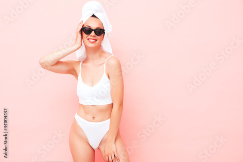 Young beautiful smiling woman in white lingerie. Sexy carefree model in underwear and towel on head posing pink wall in studio. Positive and happy female enjoying morning in sunglasses