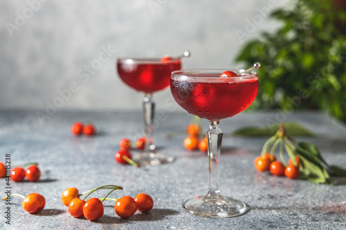 Champagne coupe glass of refreshing cherry or merry cocktail with ice served on gray table surface surround of fresh merry and different green plants, shallow depth of the field.