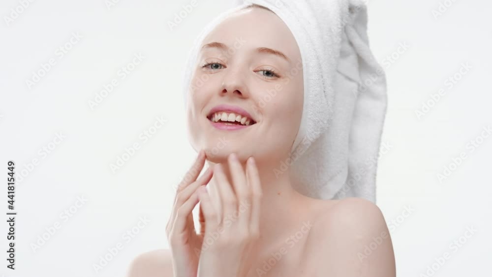 Beautiful slim European young woman with a towel on her hair touches her face smiling wide for the camera on white background | Face skin health concept Beautiful slim European young woman with a towel on her hair touches her face smiling wide for the camera on white background | Face skin health concept