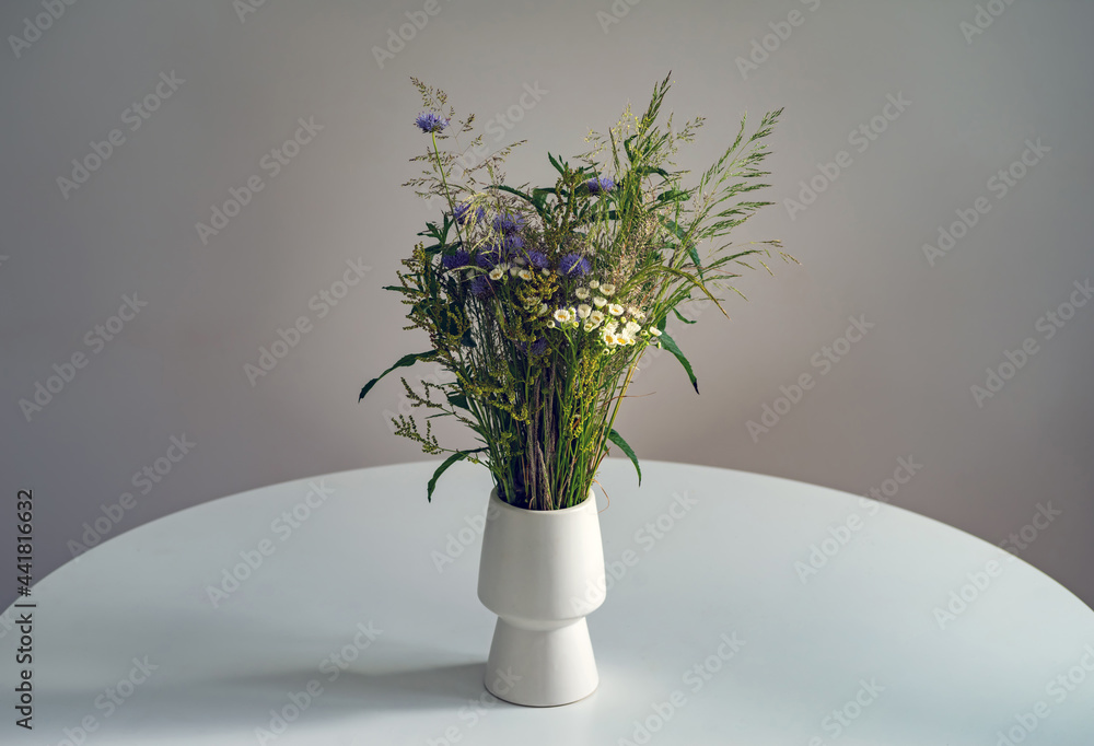 A bouquet of wild flowers in a white vase on a table on a gray background. A bouquet of wild flowers in a white vase on a table on a gray background.