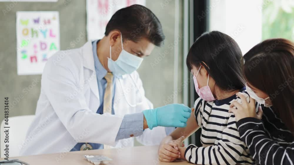 Asian senior doctor wearing gloves and isolation mask is making a COVID-19 vaccination in the shoulder of child patient with her mother at hospital. Asian senior doctor wearing gloves and isolation mask is making a COVID-19 vaccination in the shoulder of child patient with her mother at hospital.
