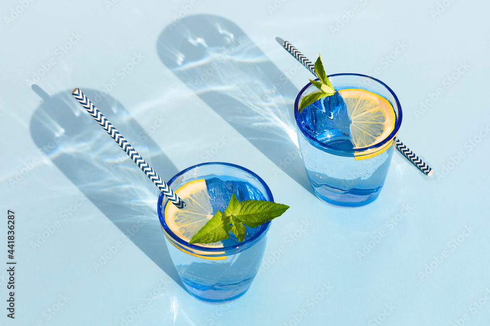 summer drink. cool refreshing beverage with mint and lemon in colored glass with paper tube. zero waste home. minimalism. natural fresh lemonade with hard shadows on blue background - obrazy, fototapety, plakaty 
