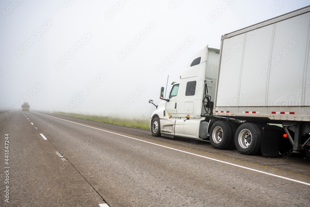 Broken white big rig semi truck with dry van semi trailer stands on the side of a foggy road waiting for mobile roadside assistance Broken white big rig semi truck with dry van semi trailer stands on the side of a foggy road waiting for mobile roadside assistance