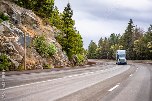 Classic gray big rig bonnet semi truck with trailer driving on the winding mountain road with rocks and trees on the sides © vit