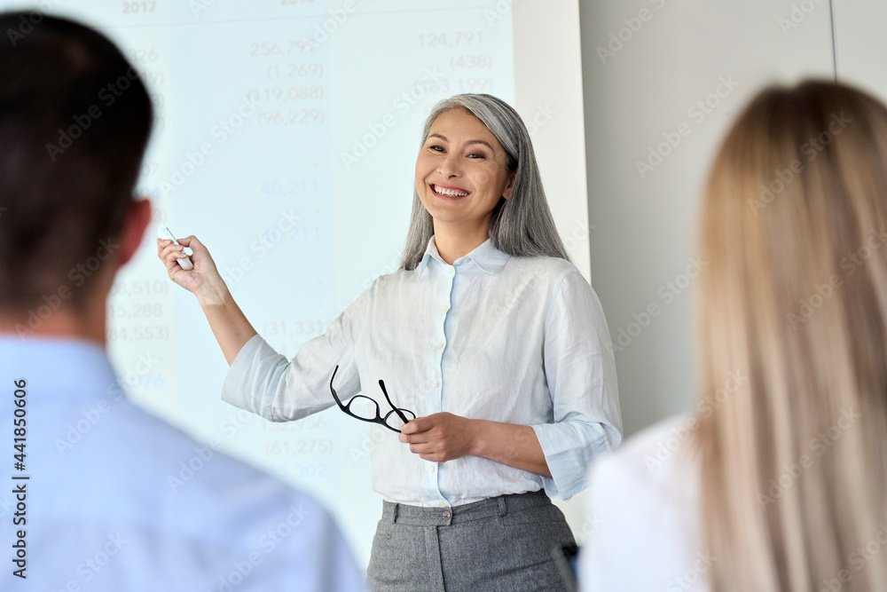 Mature happy smiling Asian corporation female executive ceo presenting project research results to interns colleagues business partners team standing pointing at board room in modern office. - obrazy, fototapety, plakaty 