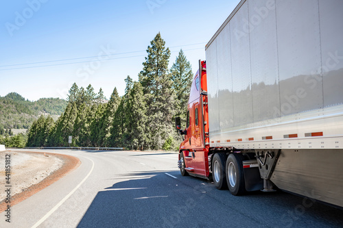 Industrial freight red big rig semi truck transporting cargo in dry van semi trailer driving downhill on the winding mountain highway road