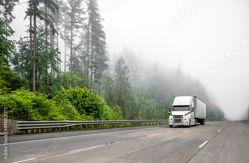 Industrial standard big rig white semi truck transporting cargo in dry van semi trailer running on the wide highway road with fog and trees.