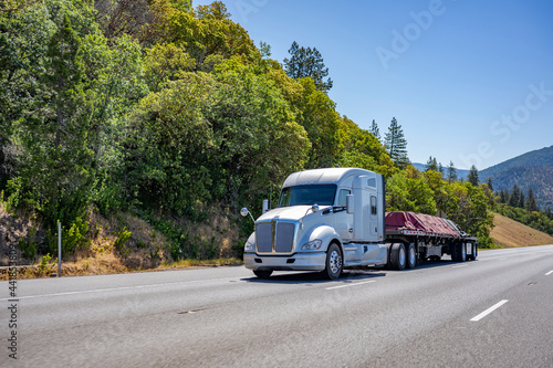 Powerful white big rig semi truck transporting tarped cargo on flat bed semi trailer climbing uphill on the wide mountain road