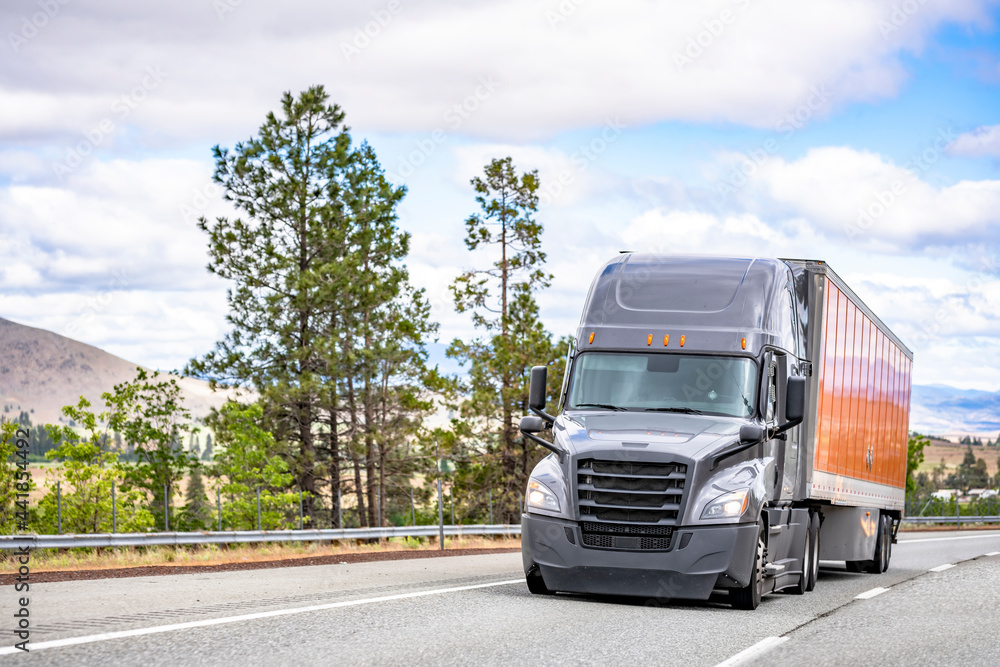 Fototapeta premium Shiny gray big rig semi truck with orange dry van semi trailer moving on the road for cargo delivery