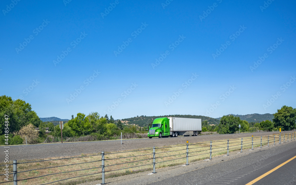 Stylish bright green big rig semi truck transporting commercial cargo in dry van semi trailer driving on the divided highway road with rope fence