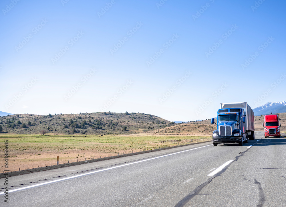 Two big rigs semi trucks red and blue colors transporting cargo in semi trailers running on the straight like arrow road