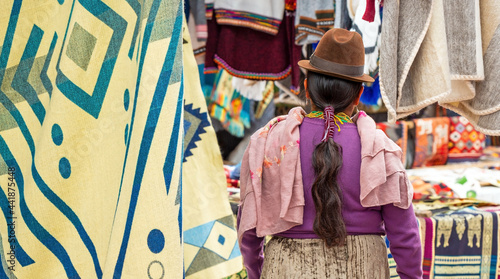 Indigenous Ecuadorian Otavalo woman in traditional clothing, hat and hairstyle on Otavalo local market with textile and fabric stalls, Ecuador. photo