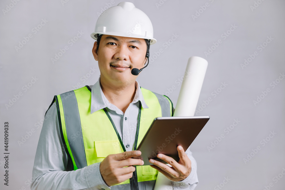 A young man, architect, or engineer wearing a reflector and a safety helmet for construction work, stands holding a design blueprint with a tablet and looking at the camera. A young man, architect, or engineer wearing a reflector and a safety helmet for construction work, stands holding a design blueprint with a tablet and looking at the camera.