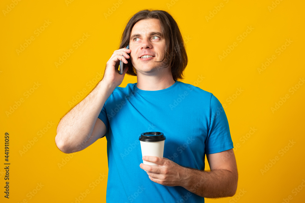 Handsome guy with funny expression, chats in cell phone, drinks hot beverage from disposable cup, dressed in casual wear.
