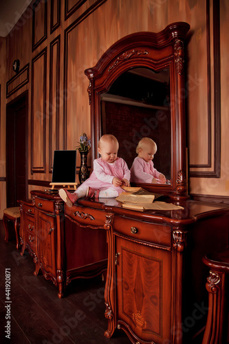 Pretty one-year-old girl in pink dress with an old book is sitting on sideboard by mirror. Child in library interior. Concept of education and child rearing. Background for site or banner. Copy space