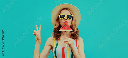 Summer portrait of stylish young woman eating a lollipop or ice cream shaped slice of watermelon wearing a straw hat on blue background © rohappy