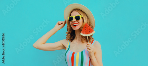 Summer portrait of happy smiling young woman with lollipop or ice cream shaped slice of watermelon wearing a straw hat on a blue background