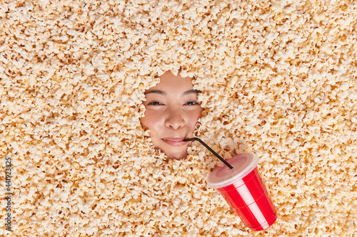 Satisfied Asian woman eats popcorn and drinks cola enjoys free time has lazy day cosumes tasty favorite snack obssesed with unhealthy food. View from above. People leisure cinematography concept photo