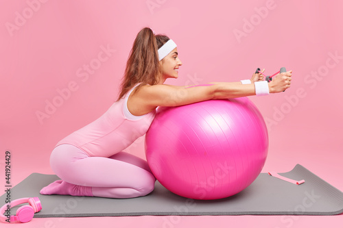 Fotografie Horizontal shot of active sli woman trains hands with expander poses on knees at fitness mat trains with swiss ball dressed in activewear isolated over pink bacground