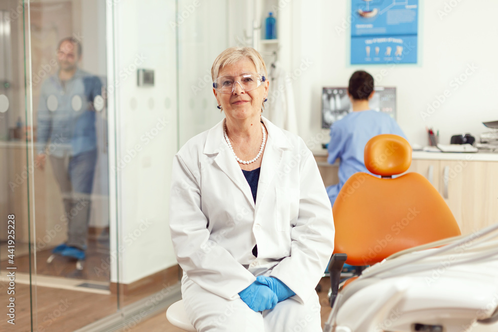 Obraz premium Orthodontist senior in medical uniform sitting on chair looking into camera waiting for man pacient to start stomatology treatment after tooth surgery. Team of dentists working in cabinet office