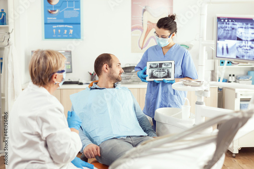 Sick man sitting on stomatological chair listening doctor while looking at tablet in dental clinic. Dentistry doctor showing to patient teeth radiography during dental consultation
