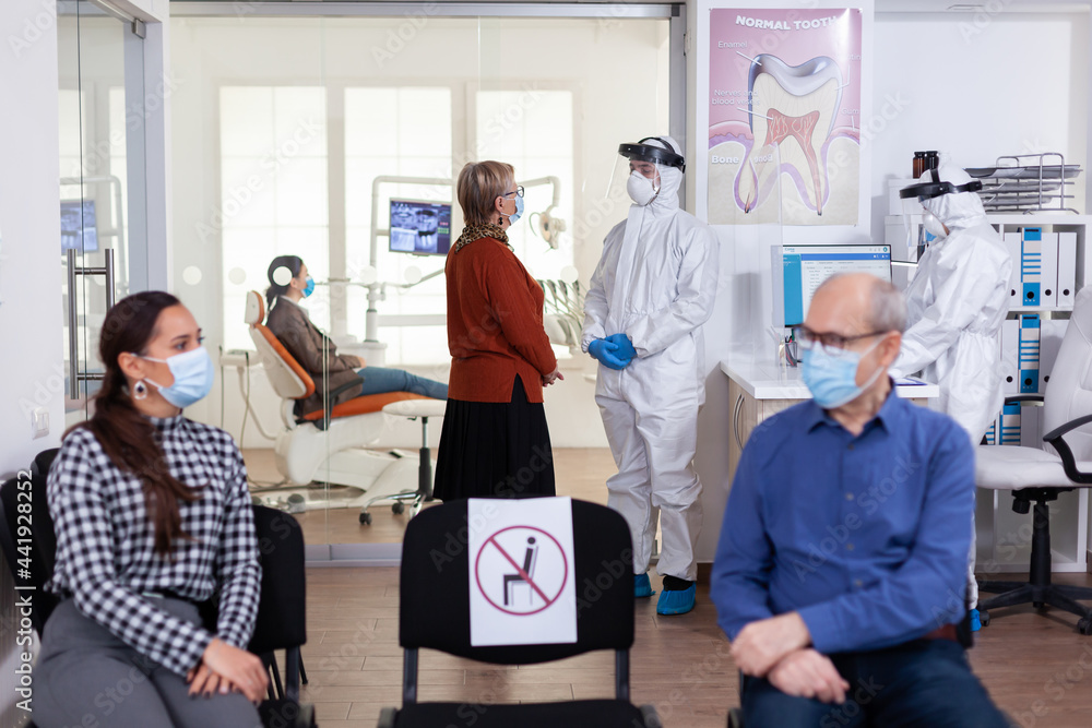 Senior woman patient in stomatology clinic discussing with doctor dressed in ppe suit as safety precaution during global pandemic with coronavirus. People talking in stomatology waiting room. Senior woman patient in stomatology clinic discussing with doctor dressed in ppe suit as safety precaution during global pandemic with coronavirus. People talking in stomatology waiting room.